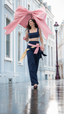 Aurelia — model walking a rain-washed street beneath an oversized pink bow umbrella, wearing a navy denim bralette and wide-leg trousers with pink and butter-yellow sash bows